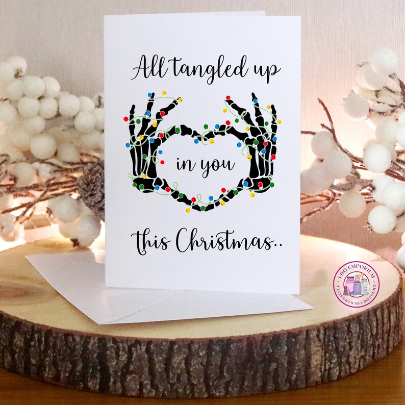 K&ouml;nnte beinhalten: Eine wei&szlig;e Gru&szlig;karte mit schwarzen Skeletth&auml;nden, die ein Herz bilden und mit bunten Lichterketten umwickelt sind. Der Text lautet: All tangled up in you this Christmas. Die Karte wird zusammen mit einem wei&szlig;en Umschlag pr&auml;sentiert.