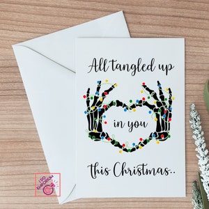K&ouml;nnte beinhalten: Eine wei&szlig;e Gru&szlig;karte mit einer Illustration schwarzer Skeletth&auml;nde, die ein Herz bilden und mit bunten Lichterketten umwickelt sind. Der Text lautet: All tangled up in you this Christmas.. Die Karte wird mit einem passenden wei&szlig;en Umschlag geliefert.