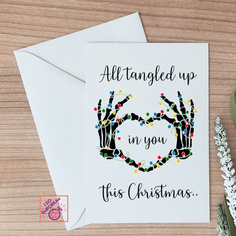 K&ouml;nnte beinhalten: Eine wei&szlig;e Gru&szlig;karte mit einer Illustration schwarzer Skeletth&auml;nde, die ein Herz bilden und mit bunten Lichterketten umwickelt sind. Der Text lautet: All tangled up in you this Christmas.. Die Karte wird mit einem passenden wei&szlig;en Umschlag geliefert.