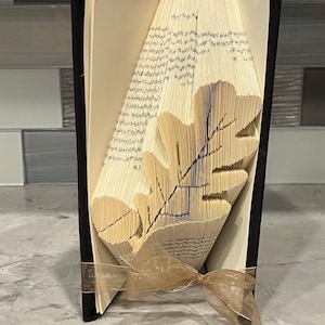 Könnte beinhalten: Eine Buchkunstskulptur eines Eichenblattes, gefaltet aus den Seiten eines Buches. Das Buch ist in schwarzem Stoff gebunden und hat ein goldenes Band darum gebunden.