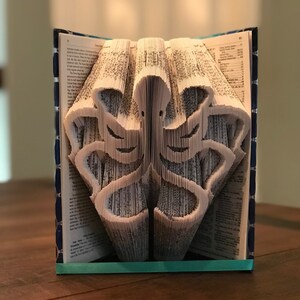 May include: A book art sculpture of an octopus made by folding the pages of a book. The sculpture is made of white paper and is sitting on a wooden table. The book is bound in blue fabric with a turquoise ribbon.