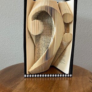 May include: A book art sculpture of a treble clef, folded from the pages of a book. The book is bound in black fabric and has a black and white checked ribbon around the bottom edge.