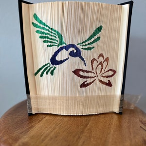 May include: A book art sculpture of a hummingbird with green wings and a blue body, hovering over a brown flower. The book is folded and bound with black fabric and a beige ribbon.