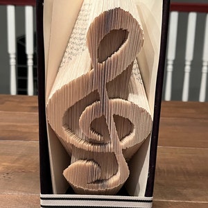 May include: A book with a black cover has been folded into the shape of a treble clef. The book is standing upright on a wooden surface. The book is decorated with a black and white ribbon.