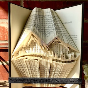 May include: A book art sculpture of a mountain range created by folding the pages of a book. The book is open and the pages are folded into a three-dimensional shape. The book is bound in black and has the word "Acknowledgments" printed on the spine.