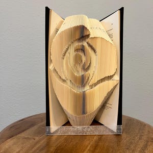 Op de afbeelding: Een boeksculptuur van een roos, gemaakt van gevouwen pagina's. De rug van het boek is zichtbaar. De sculptuur is lichtbeige en staat op een houten oppervlak.