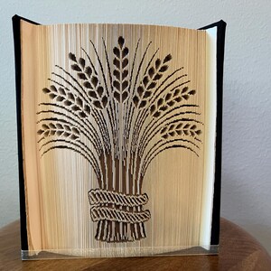 Peut inclure: Une sculpture en livre avec un design de pages pliées représentant une gerbe de blé attachée par une corde. Les pages sont de couleur crème avec des ombres sombres formant l'image, et le livre est relié par des couvertures noires.