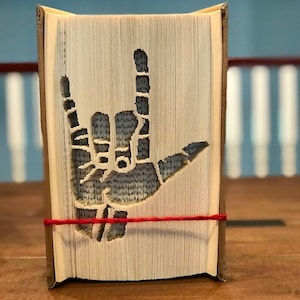 Pode incluir: Um livro com páginas dobradas para criar um gesto com a mão, com os dedos indicador e mindinho estendidos. O livro é encadernado com uma corda vermelha. O fundo é azul e branco desfocado.