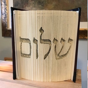 May include: A book with the Hebrew word "Shalom" cut out of the pages. The book is standing upright on a wooden surface.