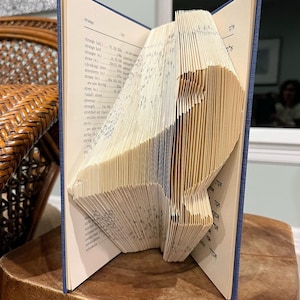 Pode incluir: Um livro dobrado na forma de um pássaro. O livro está aberto e as páginas estão dobradas para criar a forma de um pássaro. O livro está sobre uma superfície de madeira.