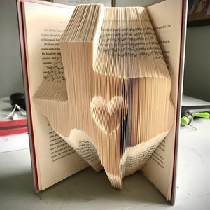 Peut inclure: Un livre plié en forme de l'état du Texas avec un cœur au centre. Le livre est ouvert et les pages sont pliées pour créer la forme.
