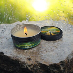 Op de afbeelding: Een brandende kaars in een zwarte blik met een groene en blauwe natuur scène en de woorden "The Scent of Rain" op de deksel. De kaars brandt met een heldere gele vlam.