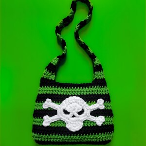 Könnte beinhalten: Gehäkelte Umhängetasche mit Totenkopf- und gekreuzten Knochen-Design. Die Tasche hat schwarze und grüne horizontale Streifen mit einer weißen Totenkopf- und gekreuzten Knochen-Applikation auf der Vorderseite. Der Riemen ist schwarz und grün.