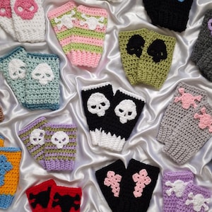 Puede incluir: Una colección de guantes sin dedos de ganchillo en varios colores, con diseños de calaveras. Los guantes son en tonos blanco, rosa, verde, azul, negro, gris, morado y rojo. Los motivos de calaveras son en colores contrastantes.