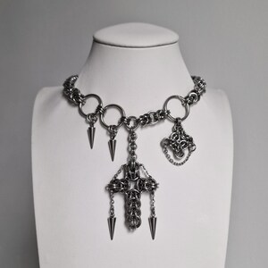 Puede incluir: Un collar de cota de malla plateado con un diseño gótico. El collar presenta una cadena tipo gargantilla, eslabones circulares grandes y elementos colgantes que incluyen tres puntas en forma de cono y un colgante de cruz. El colgante de cruz tiene tres puntas en forma de cono adicionales.