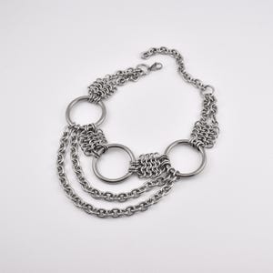 Puede incluir: Un collar plateado con tres grandes anillos circulares conectados por cota de malla y eslabones de cadena. El collar tiene tres hilos de cadena que cuelgan del anillo central. El collar está sobre un fondo blanco.