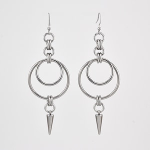 Puede incluir: Un par de pendientes plateados. Cada pendiente presenta una serie de anillos conectados, incluyendo dos aros grandes y anillos de conexión más pequeños. Un dije cónico y puntiagudo cuelga de la parte inferior. Los pendientes tienen un diseño moderno y geométrico.