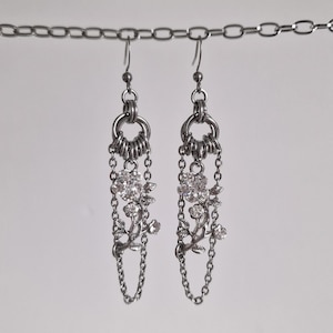 Puede incluir: Un par de pendientes colgantes plateados. Cada pendiente presenta un diseño floral con cristales transparentes y detalles de cadena. Los pendientes cuelgan de una cadena.