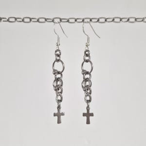 Peut inclure: Une paire de boucles d'oreilles pendantes argentées. Chaque boucle d'oreille présente une série d'anneaux et de chaînes reliés, se terminant par un petit pendentif en forme de croix. Les boucles d'oreilles sont suspendues à un crochet argenté.