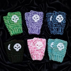 Puede incluir: Seis guantes sin dedos de ganchillo en varios colores: verde, morado, azul, verde oscuro, rosa y turquesa. Cada guante tiene una calavera blanca. Se muestran sobre un fondo negro texturizado.