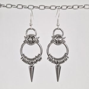 May include: A pair of silver-toned earrings with a unique design. Each earring features a circular hoop with coiled wire, a central woven element, and a conical spike pendant. The earrings are suspended from a chain.