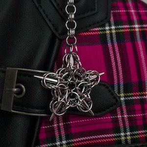 Może przedstawiać: Srebrny urok w kształcie gwiazdy z kolczugi z ostrymi końcówkami wisi na srebrnym łańcuszku. Urok jest przymocowany do czarnego skórzanego paska ze srebrną klamrą. Różowo-czarny wzór w kratę jest widoczny w tle.