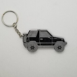 Könnte beinhalten: Ein schwarzer und grauer Schlüsselanhänger mit dem Seitenprofil eines kleinen SUVs. Der Schlüsselanhänger hat einen silbernen Metallring.