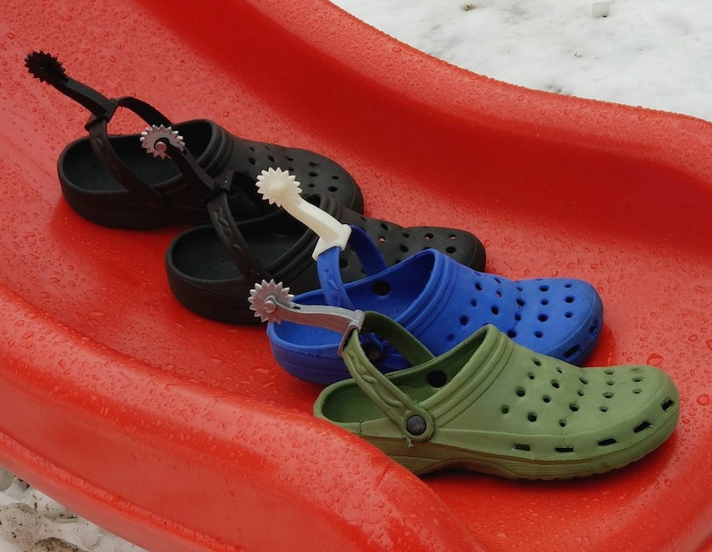 May include: Three pairs of clogs with a cowboy boot spur attached to the strap. The clogs are black, blue, and green.