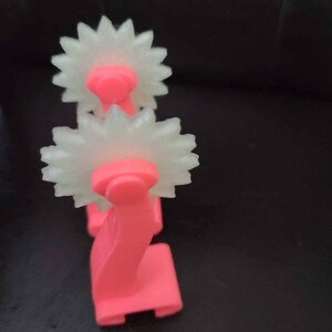 May include: A pink plastic tool with two white, star-shaped wheels. The tool is used for creating decorative patterns in clay or fondant.