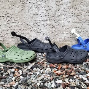 May include: Three pairs of green, black, and blue clogs with silver, black, and white spur attachments. The clogs are on a bed of small gray rocks.