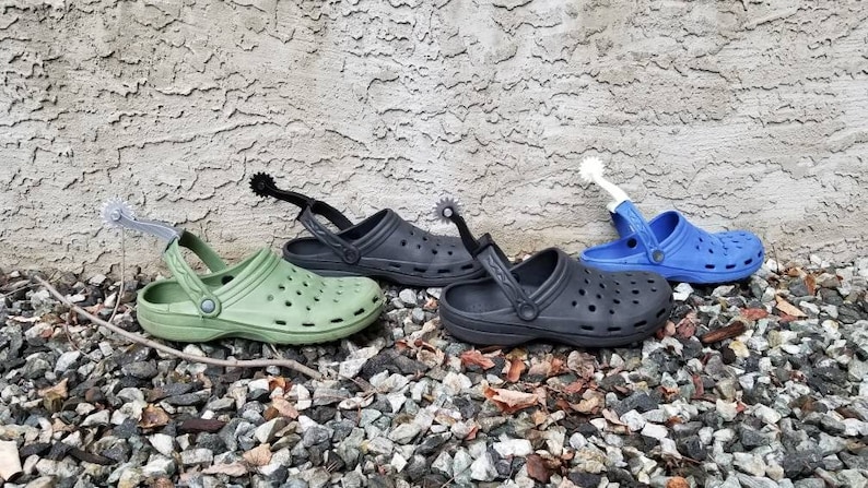 May include: Three pairs of green, black, and blue clogs with silver, black, and white spur attachments. The clogs are on a bed of small gray rocks.