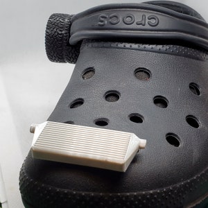 May include: A black Crocs clog with the word "Crocs" printed on the strap. A white plastic object is sitting on top of the clog.