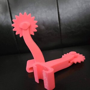 May include: Two pink plastic spur-shaped objects with a star-shaped design on the end. The spurs are connected by a metal bar.