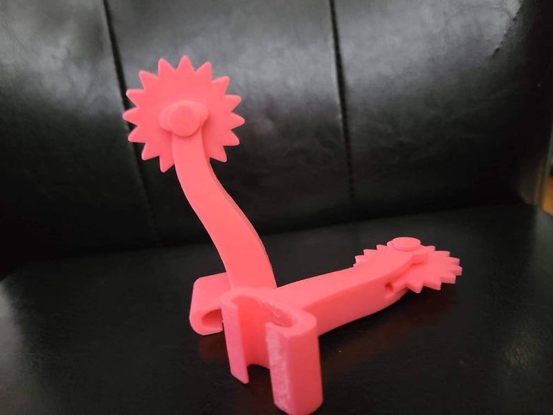May include: Two pink plastic spur-shaped objects with a star-shaped design on the end. The spurs are connected by a metal bar.