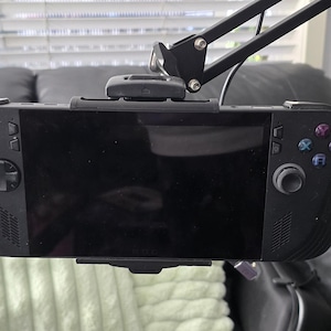 May include: Black handheld gaming console with a large screen, mounted on a black adjustable arm. The console has control buttons on both sides. The background includes a sofa, a green pillow, and a decorative lamp.