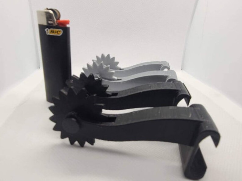 May include: Three 3D printed black and gray gear-shaped objects with a black lighter in the background.