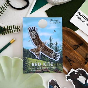 May include: A rectangular product card featuring a red kite bird illustration in flight over a green landscape. The text reads Ferne Creative, Red Kite, Illustrated Iron-On Patch, Designed in the UK, Made in Small Batches. The background is blue sky.