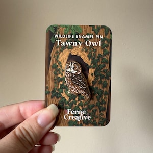May include: A Tawny Owl enamel pin attached to a rectangular backing card. The card features an illustration of a tree trunk with green ivy and the text Wildlife Enamel Pin, Tawny Owl, and Ferne Creative. The pin depicts a brown owl.