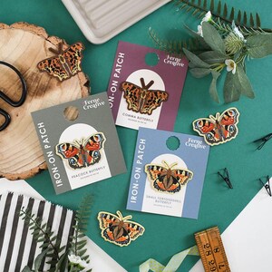 May include: A collection of butterfly iron-on patches, each with intricate designs and vibrant colours. The patches are presented on cards, with the text "IRON-ON PATCH" and butterfly names. The brand name "Ferne Creative" is visible. The background is a teal green.