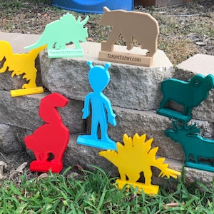 May include: A set of 10 colorful 3D printed animal and creature targets for shooting practice. The targets include a dinosaur, an alien, a bear, a ram, a moose, and a triceratops. The targets are all on a stone wall background. The text "TargetSafari.com" is printed on the base of each target.