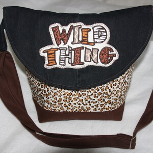 Könnte beinhalten: Eine braune und schwarze Umhängetasche mit einer Klappe, auf der ein weißer Patch mit den Worten "WILD THING" in bunten Fäden gestickt ist. Die Tasche hat ein Leopardenmuster-Futter.