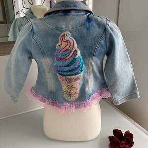 Könnte beinhalten: Eine hellblaue Jeansjacke mit rosa Rüschenbesatz und einem Pailletten-Eiswaffendesign auf der Rückseite.