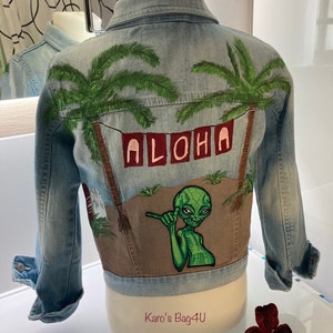 Könnte beinhalten: Eine hellblaue Jeansjacke mit einem bemalten Design auf der Rückseite. Das Design zeigt Palmen, das Wort "ALOHA" in roten und rosafarbenen Buchstaben und einen grünen Außerirdischen mit einer Handgeste. Die Jacke befindet sich auf einem weißen Mannequin.