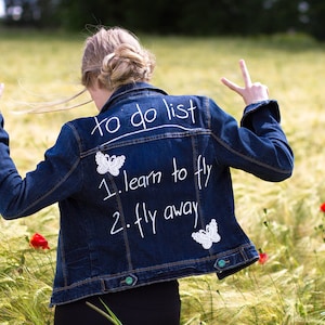 Könnte beinhalten: Eine blaue Jeansjacke mit weißer Stickerei, auf der "to do list 1. learn to fly 2. fly away" steht. Die Jacke hat zwei weiße Schmetterlingsapplikationen auf der Rückseite.