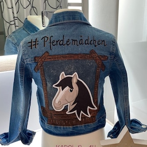 Könnte beinhalten: Eine blaue Jeansjacke mit einem braunen Holzrahmen und einer Pferdekopf-Illustration. Der Text "#Pferdemädchen" ist auf der Rückseite der Jacke gestickt.