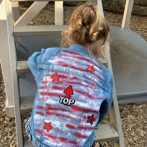 Könnte beinhalten: Eine blaue Jeansjacke mit roten und weißen Streifen und roten Sternen. Die Jacke hat den Schriftzug "TOP" mit einem nach oben zeigenden Pfeil.