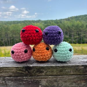 May include: Six colourful, crocheted spheres stacked on a wooden surface. The spheres are red, purple, pink, orange, and mint green, each with two black eyes. The background features a blurred view of a green field and trees under a blue sky.