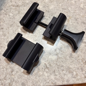 May include: Two black plastic mounting clamps with a matte finish. One clamp features an adjustable threaded metal screw mechanism with a flared handle for tightening, while the other is a separate, matching component with a grooved channel.