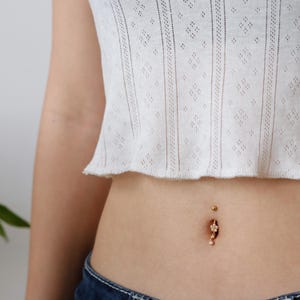May include: A gold-coloured belly button ring with a floral design. The ring features small, clear gemstones and hangs below the navel. The person is wearing a white, open-weave crop top.