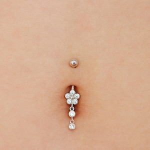 May include: A silver belly button ring featuring a flower design and clear gemstones. The flower has six petals, each set with a small, clear stone. Two additional clear gemstones dangle below the flower.